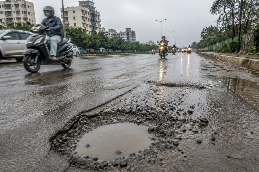 A rain-wet road with a large pothole on a Bengaluru street in Varthur, showing damaged urban road infrastructure