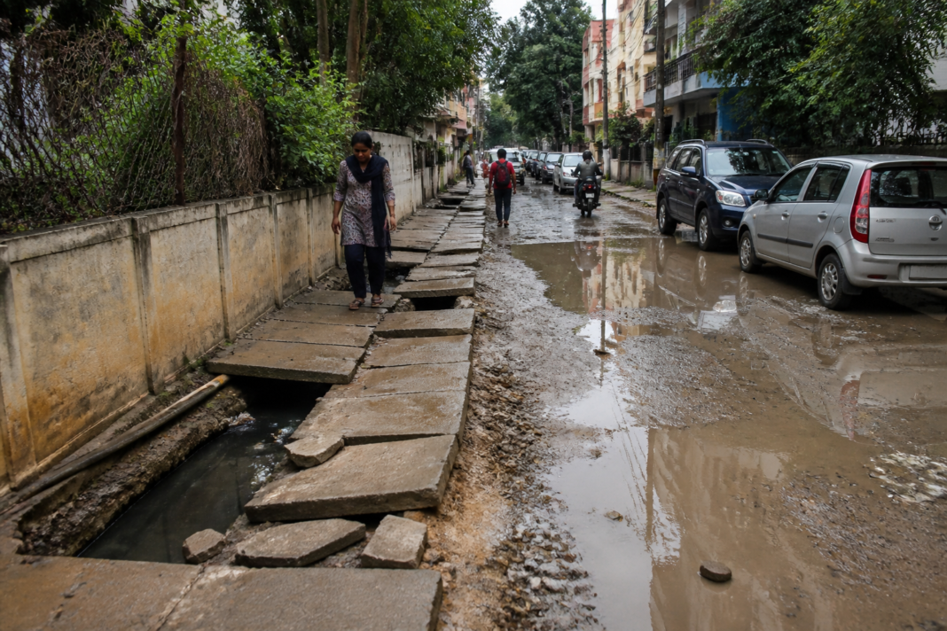 A damaged footpath beside an open drain and muddy road in a Bengaluru neighbourhood