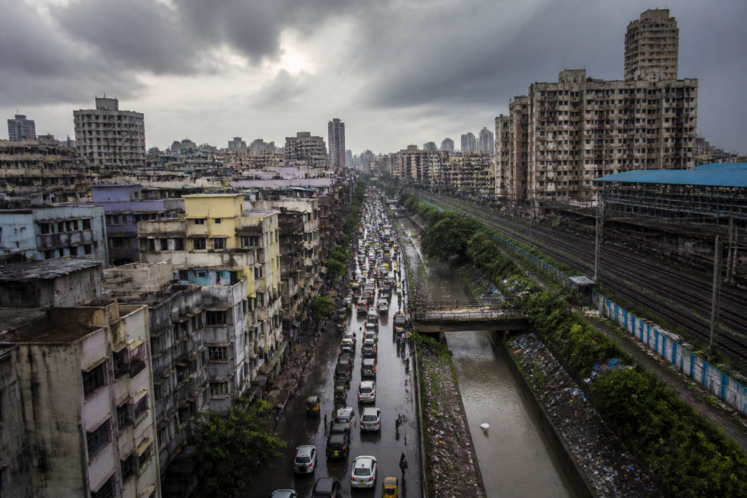 Dense Indian city streets and buildings under heavy monsoon clouds, illustrating the urban crisis and pressure on infrastructure