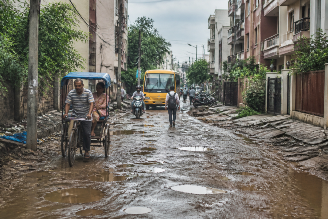 Aali Vihar road showing residents and vehicles using an uneven unpaved route in Delhi.