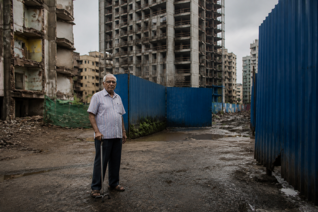 Mumbai redevelopment delays leave elderly flat owners waiting outside unfinished housing projects.