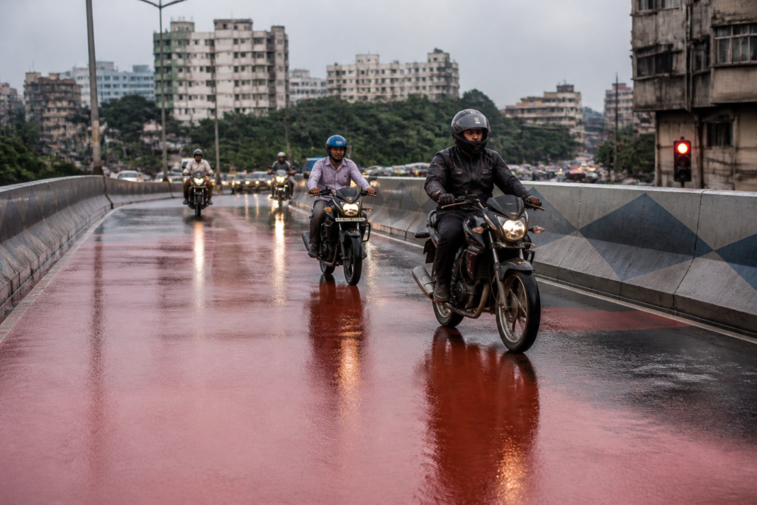 Wet Kolkata flyover surface after rain, with motorbikes navigating the coloured road coating.
